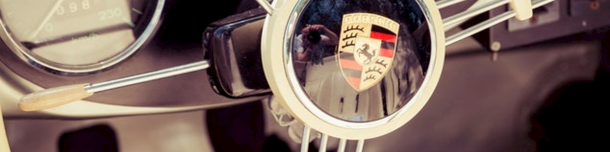 The image shows the dashboard and steering wheel of a classic car, featuring the Porsche logo at the center of the steering wheel.