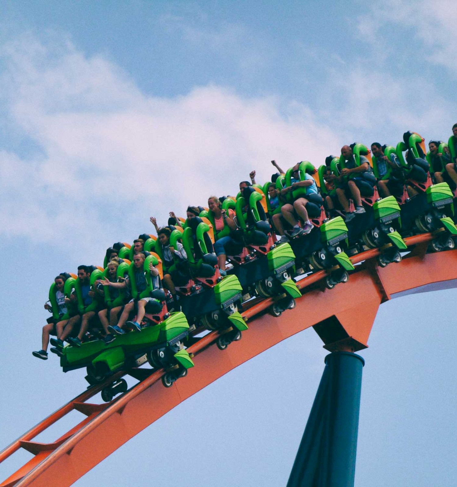 People are riding a green and orange roller coaster, ascending a steep track against a blue sky with some clouds.