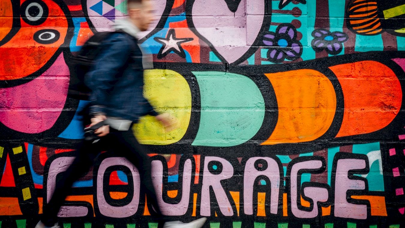 A person walks by a colorful graffiti mural featuring abstract shapes, hearts, stars, and the word "COURAGE" in bold letters.