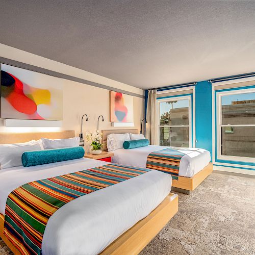 A modern hotel room features two beds with colorful striped runners, wall art, large windows, and stylish furnishings.