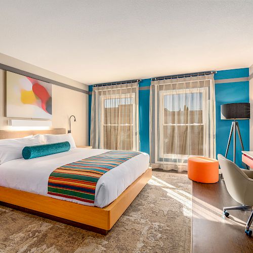 A modern hotel room with a king-size bed, colorful decor, large windows, a desk with a chair, a floor lamp, and an orange ottoman.