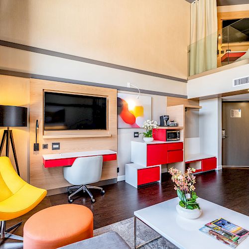 A modern loft-style living space with a TV, desk, chair, bright yellow armchair, staircase, and a mezzanine level.