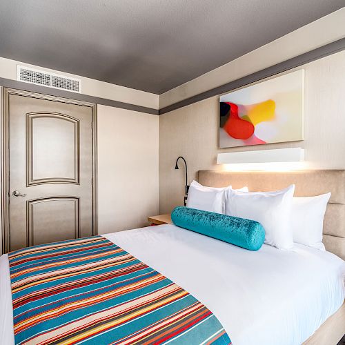 A modern hotel room with a colorful bed runner, teal bolster pillow, abstract wall art, and bedside tables with mounted lamps ends the sentence.