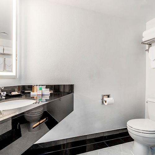 A modern bathroom with a sink, a mirror, a toilet, toiletries on the counter, and towels hanging on a rack.