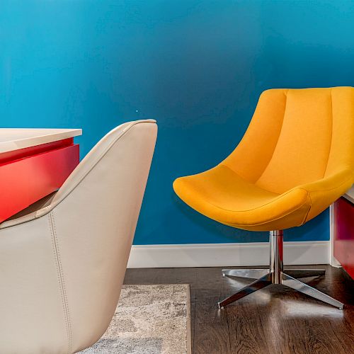 The image features a modern office setup with a white chair, red desk, blue wall, and a vibrant yellow chair with a metal base, on wooden flooring.