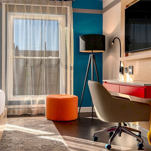 A modern bedroom with a blue and beige color scheme, featuring a bed, window with sheer curtains, desk, chair, TV, floor lamp, and an orange ottoman.