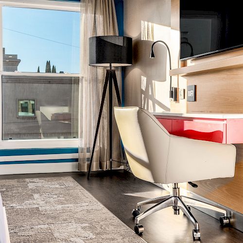 A well-lit modern room with a desk, chair, TV, standing lamp, and window with curtains. The desk is red and white, and the floor is carpeted.