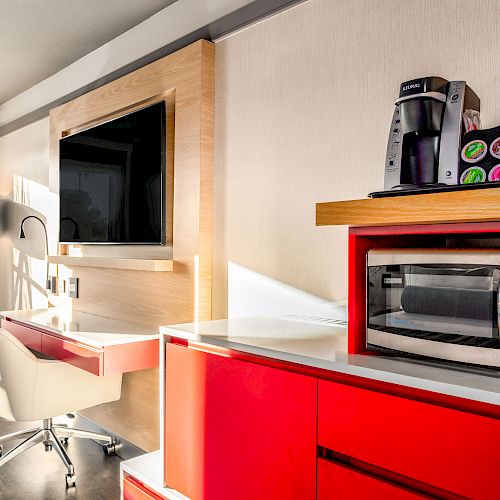 A hotel room's workstation with a white chair, desk, TV, lamp, curtains, microwave, coffee maker with pods on a red cabinet, and window.