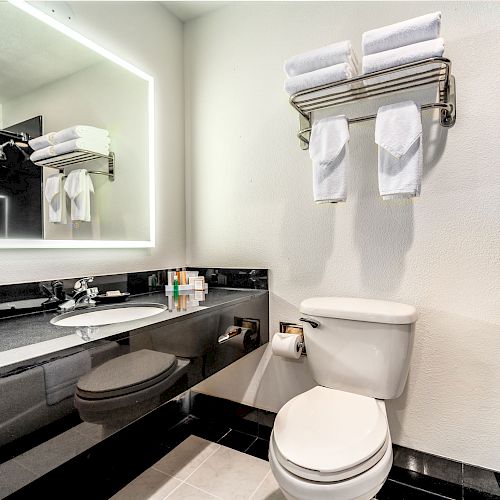 A modern bathroom features a backlit mirror, sink with toiletries, toilet, and a towel rack with neatly folded towels on the wall.