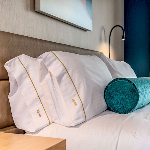 The image shows a neatly made bed with white pillows featuring gold accents, a teal cylindrical cushion, and a wall-mounted reading lamp next to it.