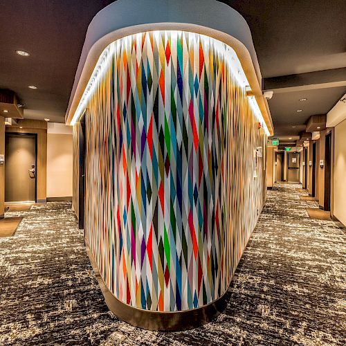 A hotel hallway with patterned carpet and a vibrant, multicolored abstract design on a rounded wall, doors to the rooms lining either side.