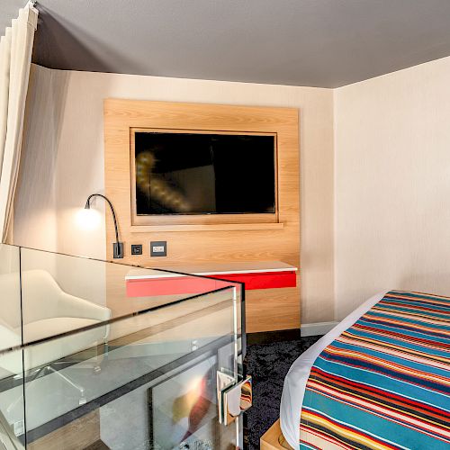 A modern hotel room with a wall-mounted TV, a colorful striped bed cover, and a glass balcony railing is shown, ending the sentence.