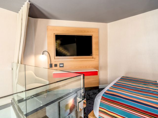 A hotel room with a glass desk, striped bed, and flat screen TV on a wooden panel.