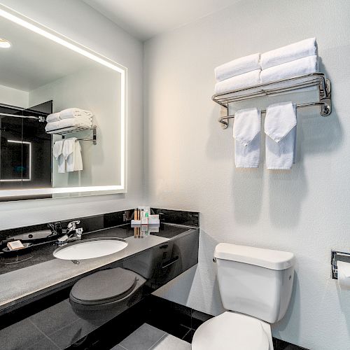 A modern bathroom with a lit mirror, sink, toiletries, toilet, and a rack with folded towels and washcloths on the wall above the toilet.