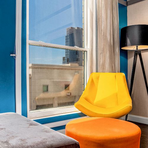 A modern room with a large window, yellow chair, orange ottoman, black floor lamp, and blue accents; cityscape in the background ends the sentence.