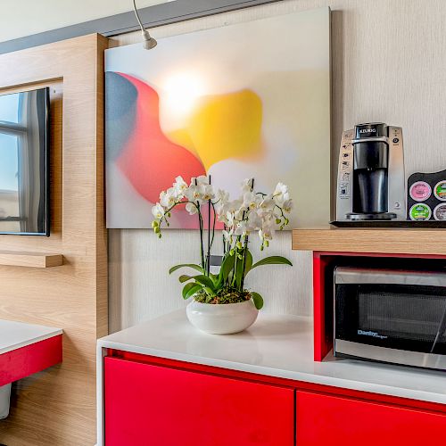 The image shows a modern interior featuring a flat-screen TV, a colorful abstract painting, a potted orchid, a coffee maker, and a microwave.