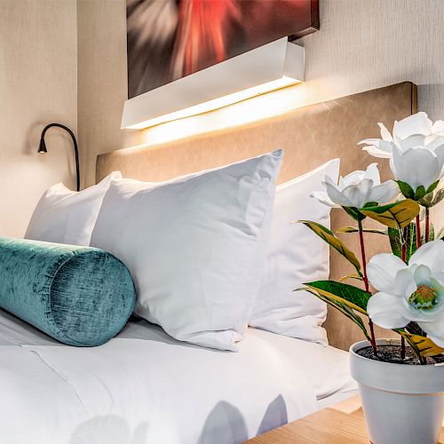 A neatly made bed with decorative pillows, a nightstand with a potted plant featuring white flowers, and a wall-mounted reading light.