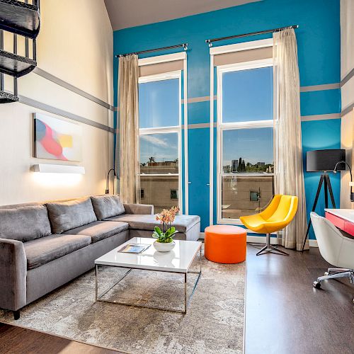 A modern living room with a gray sofa, colorful chairs, a desk, a TV, and a spiral staircase, plus large windows with a city view and bright accents.