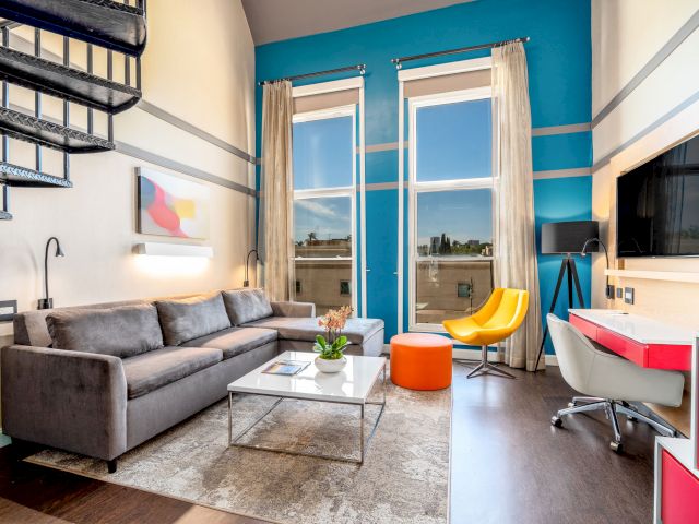 A wide view of a living room with a gray sofa, white coffee table, and desk area, featuring a vibrant blue accent wall and large windows with a city view.