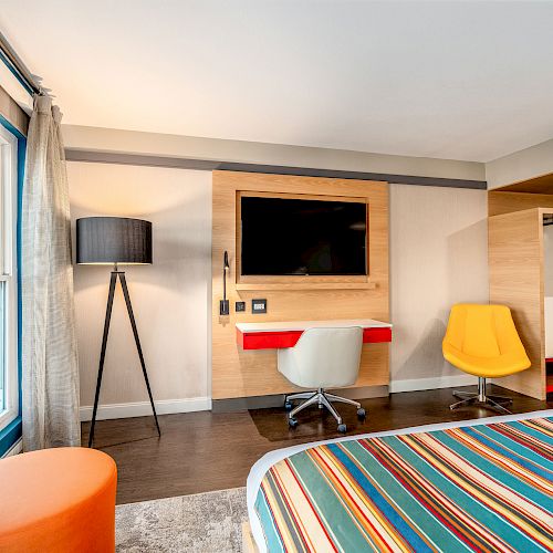 A modern hotel room features a bed with colorful striped linens, a wall-mounted TV, a desk, two chairs, a floor lamp, and a large window.