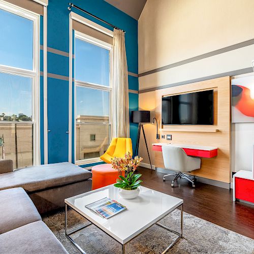 A modern living room with a sectional sofa, wall-mounted TV, desk, bright accent chairs, and large windows flooding the space with natural light.