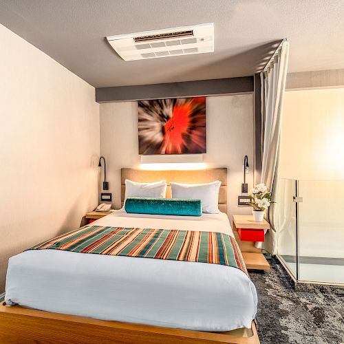 A modern bedroom with a colorful bedspread, abstract art above the bed, glass railings, and hanging lights, creating a stylish and cozy atmosphere.