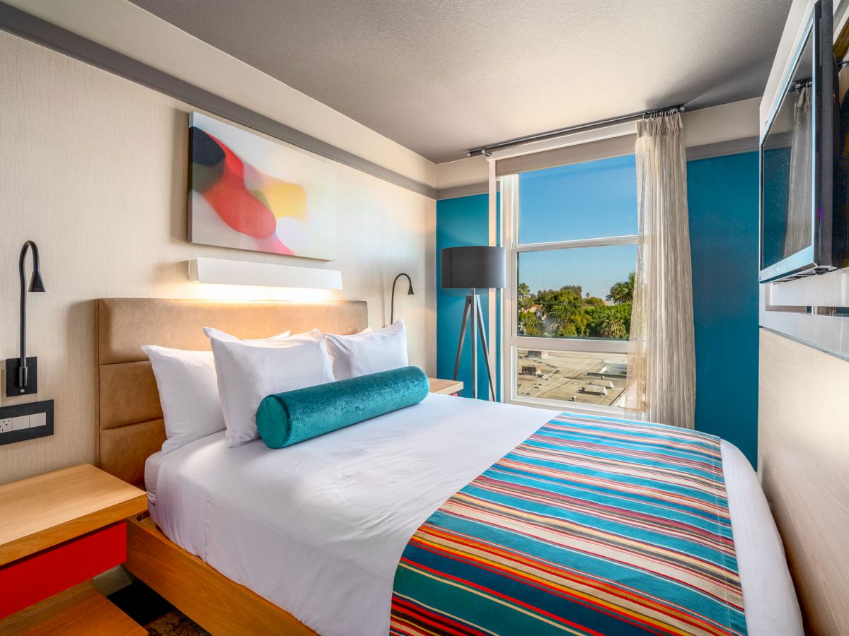 A modern bedroom with a neatly made bed, colorful bedding, abstract wall art, bedside tables, and a window overlooking an outdoor view.