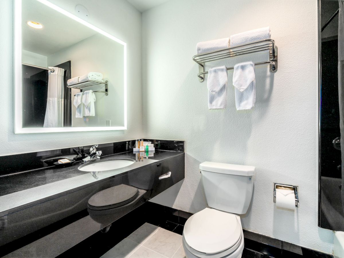 Modern bathroom with a lit mirror, sink, toiletries, toilet, towel rack with folded towels, and a shower curtain in the background.