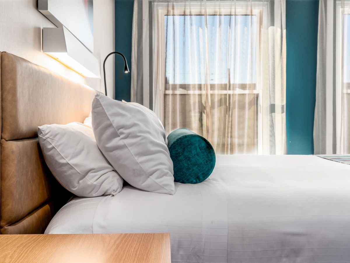 A neatly made bed with white linens and pillows, a teal bolster cushion, and two large windows covered with sheer curtains.