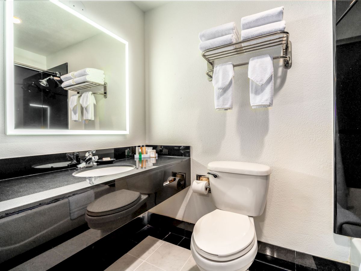 The image shows a modern bathroom with a toilet, a sink, a large mirror, and towels neatly arranged on a shelf above the toilet.