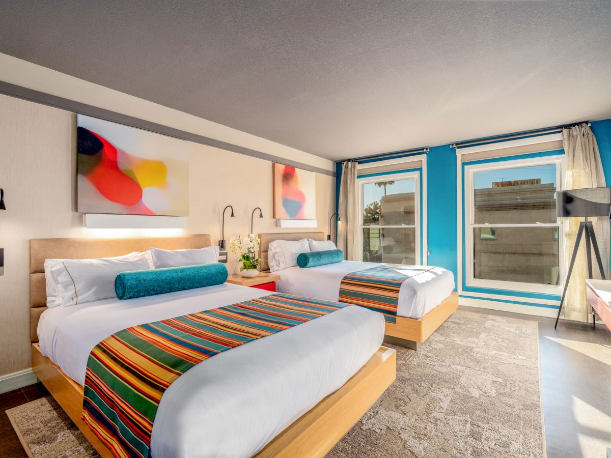 A modern hotel room with two beds featuring colorful striped runners, wall art, tall windows, and contemporary lighting.