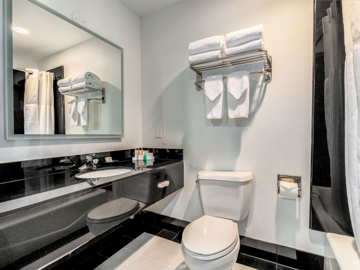 A bathroom with a large mirror, black countertop, sink, and a toilet. Towels are neatly stacked and hanging above the toilet.