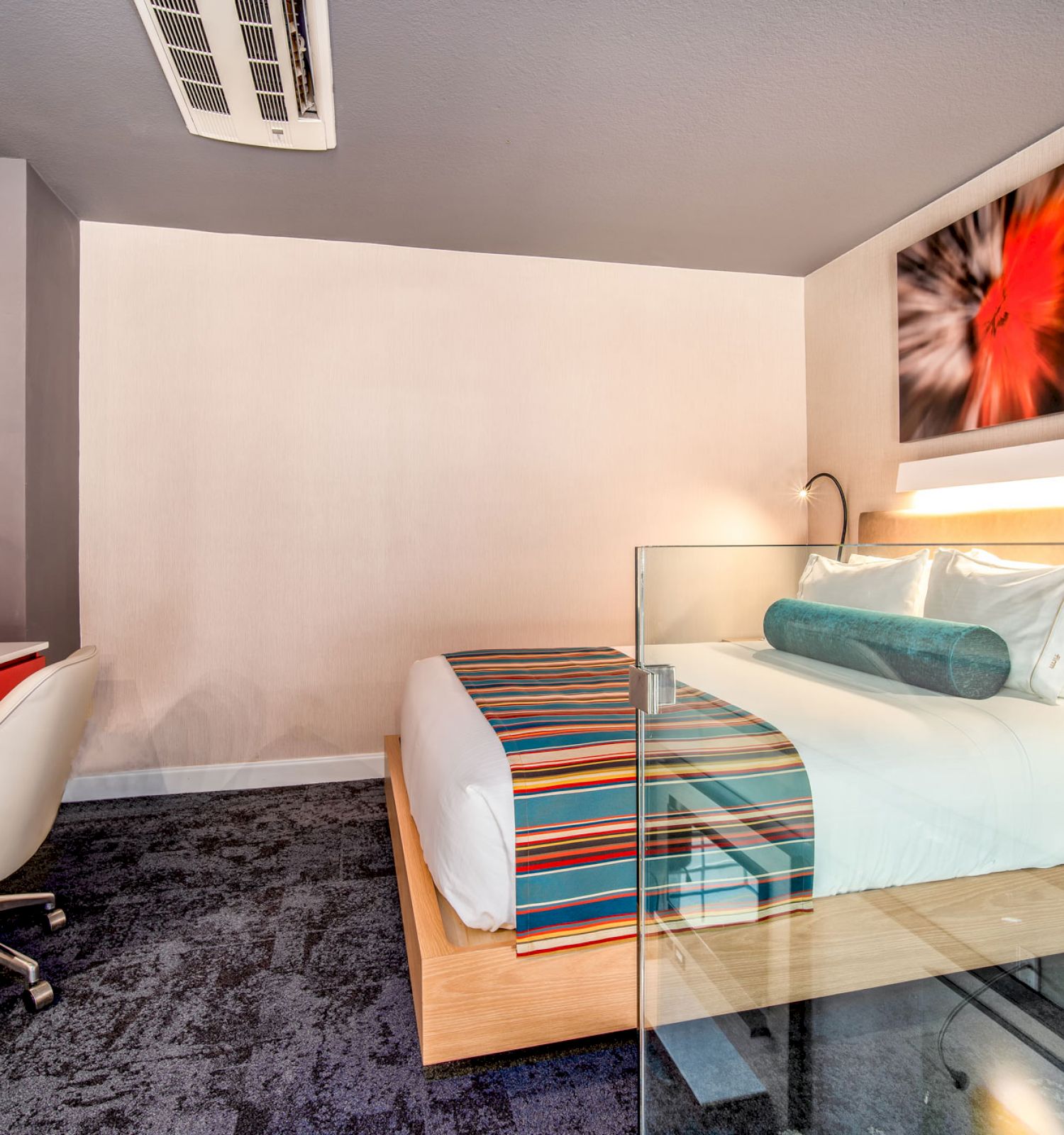 The image shows a modern hotel room with a bed, vibrant bedding, a large wall-mounted TV, a desk with a chair, and a piece of abstract artwork above the bed.