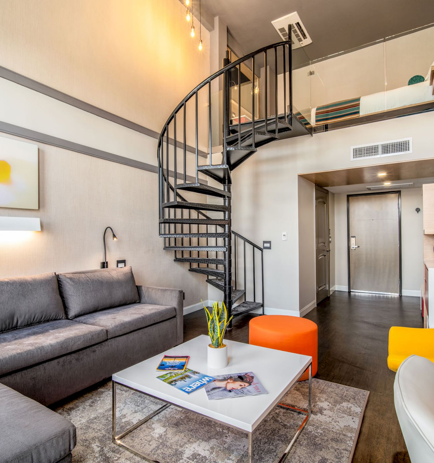 A modern loft apartment with a spiral staircase, colorful furniture, gray sofa, and a white coffee table with magazines and a plant.