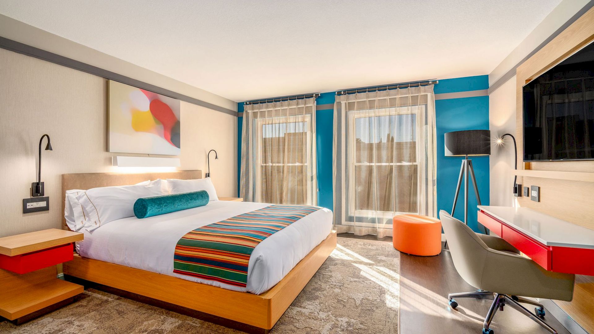 A modern hotel room with a king-sized bed, abstract wall art, a work desk, a chair, an orange stool, and large windows with sheer curtains.