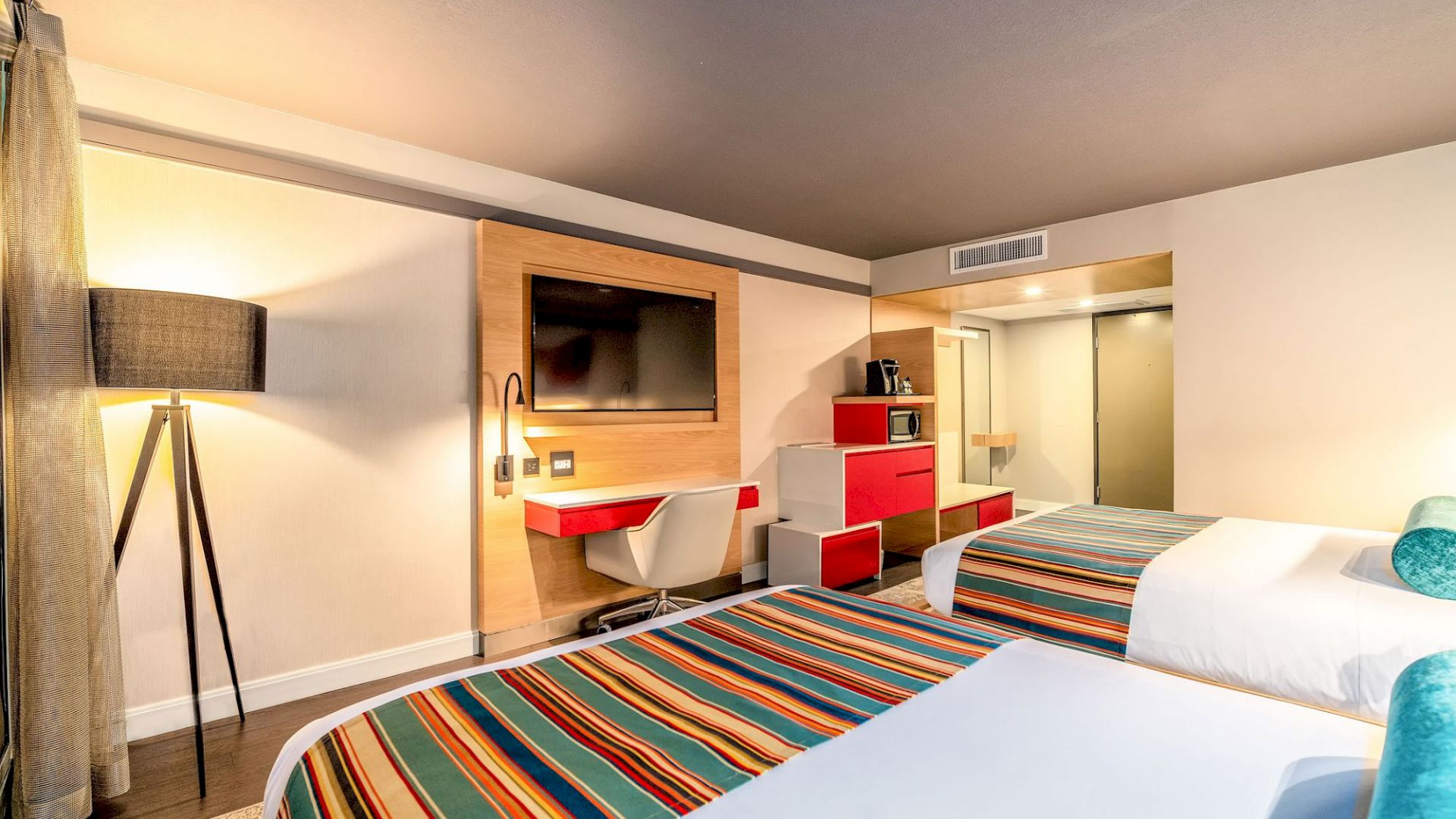 A modern hotel room with two beds, striped bedding, a flat-screen TV, a desk with a chair, and a floor lamp, featuring a bright, functional design.