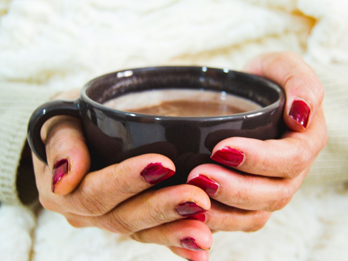 A pair of hands with red nail polish is holding a black mug filled with a hot beverage, possibly coffee or hot chocolate, against a cozy background.