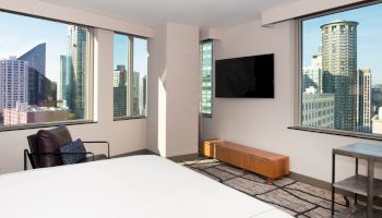 A modern room with large windows offers a cityscape view, a wall-mounted TV, a chair, a wooden cabinet, and a bed in the foreground.