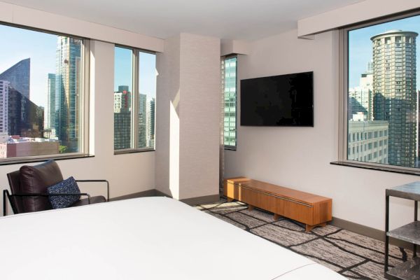 A hotel room with a city view, featuring a bed, chair, TV, and wooden furniture.