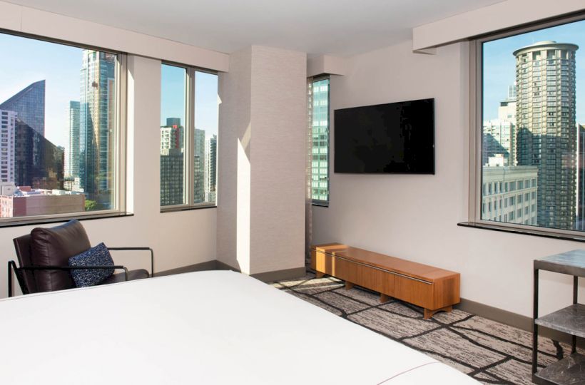 A modern room with large windows offers a cityscape view, a wall-mounted TV, a chair, a wooden cabinet, and a bed in the foreground.