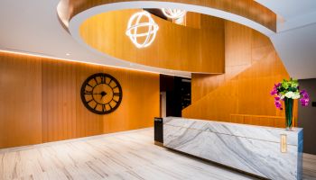 This image depicts a modern lobby area with wooden paneling, a large decorative wall clock, a marble reception desk, and stylish lighting fixtures.