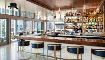 This image shows a modern bar with high shelves filled with various bottles, a wooden counter, bar stools, and large windows overlooking a cityscape.