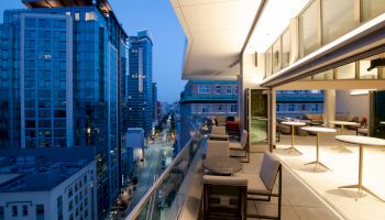 A rooftop terrace with outdoor seating overlooks a city street lined with modern buildings and skyscrapers, under a twilight sky.