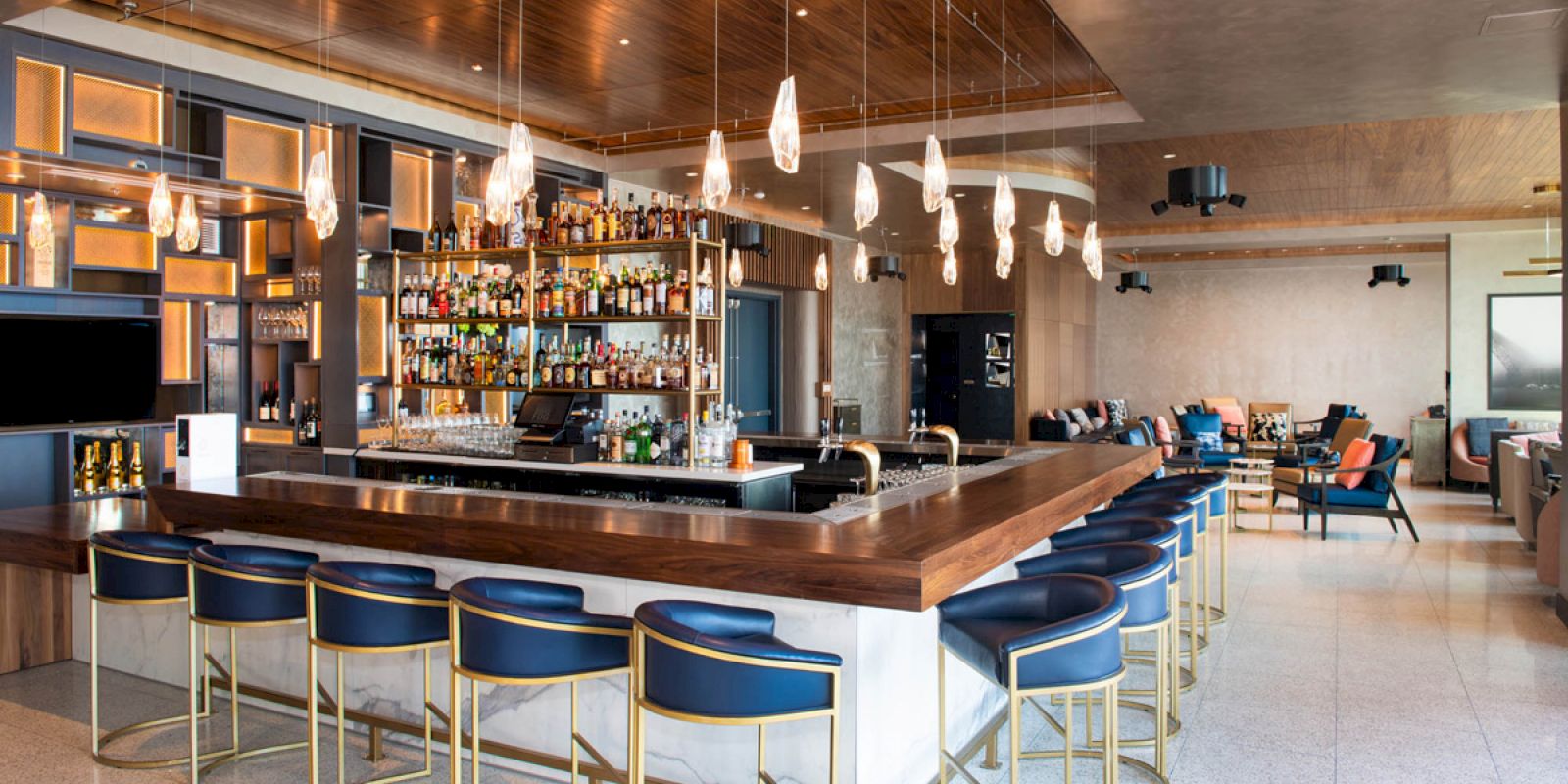 An upscale bar with a central counter, blue cushioned bar stools, hanging lights, and an extensive display of bottles. The background has seating areas.