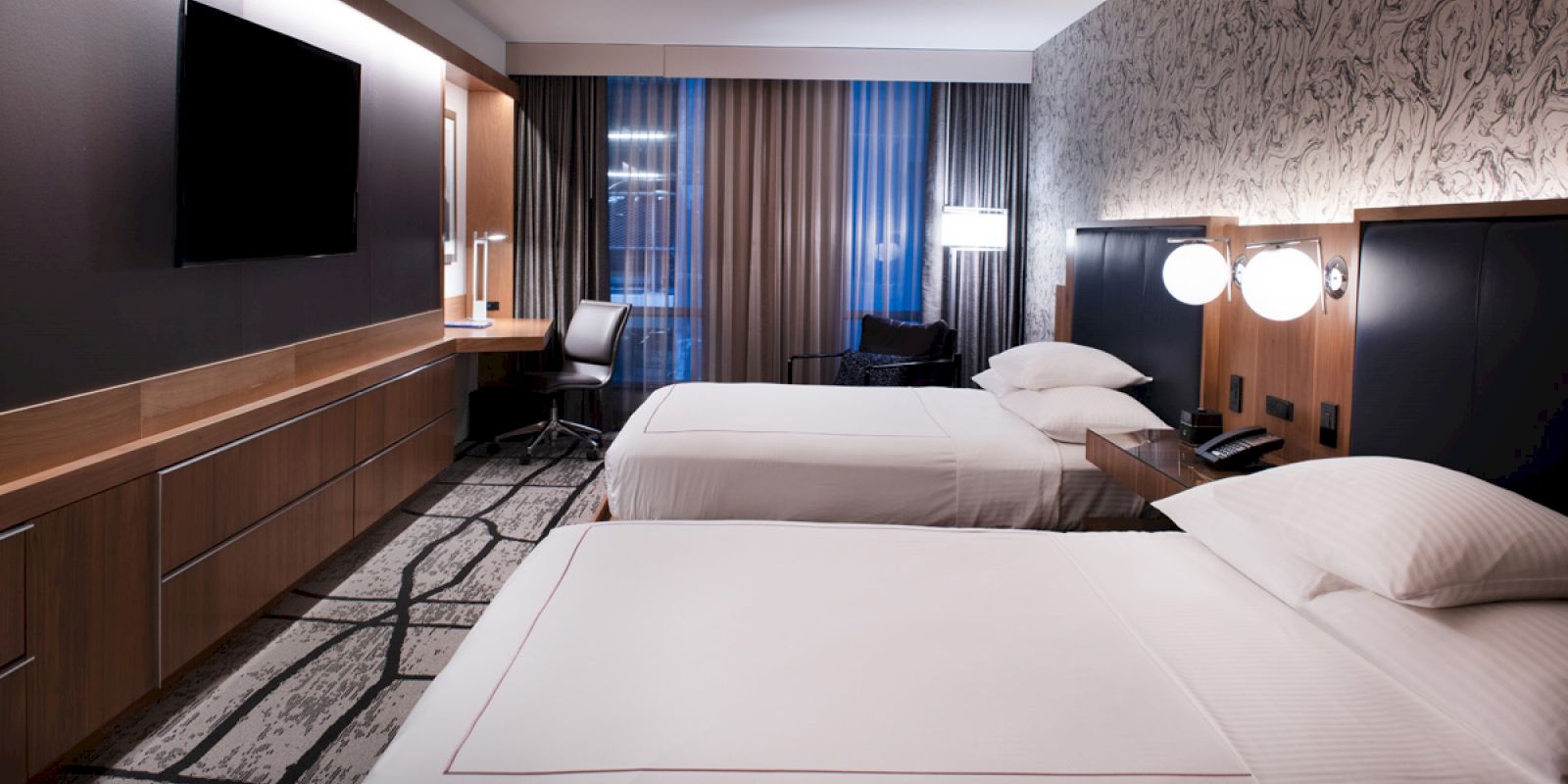 A modern hotel room with two neatly made beds, a large flat-screen TV, a work desk and chair, and stylish lighting.