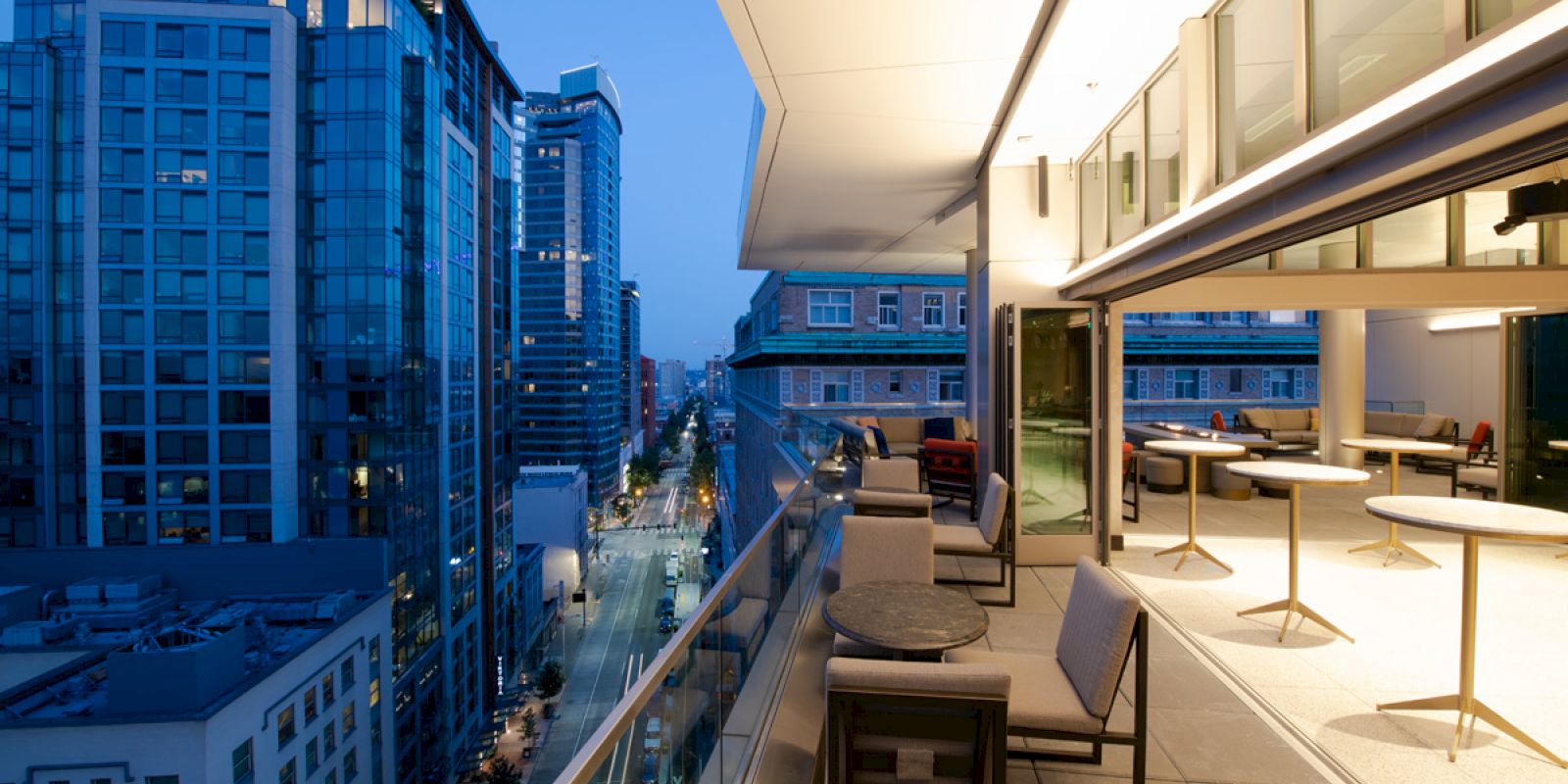 The image shows a rooftop patio with chairs and tables, overlooking a cityscape with tall buildings and a street below during dusk.