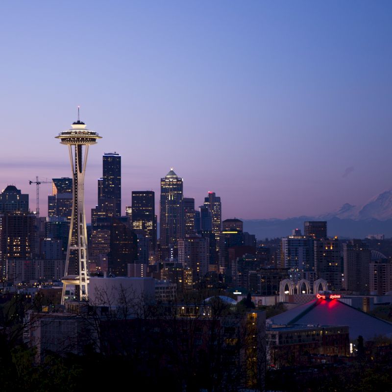This image shows the Seattle skyline at dusk, featuring the Space Needle, various skyscrapers, and a distant view of Mount Rainier.