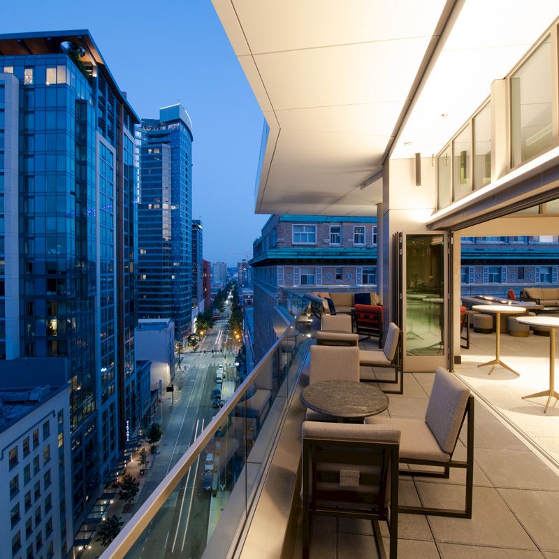 A modern cityscape view from a high-rise balcony at dusk, featuring outdoor seating and tables overlooking illuminated buildings and a street.
