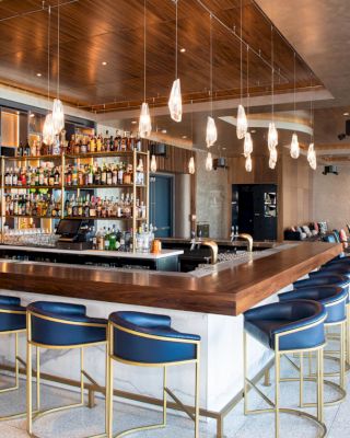 The image shows a modern, stylish bar with blue chairs, hanging lights, and a well-stocked display of bottles. There is seating in the background.