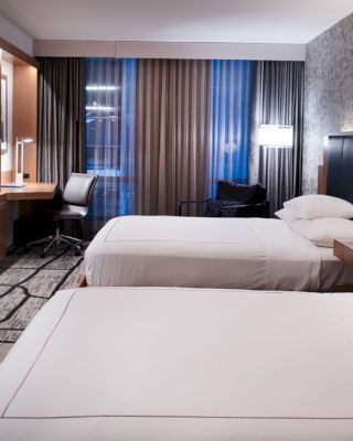 The image shows a modern hotel room with two neatly made double beds, a desk, a flat-screen TV, and a large window with curtains.